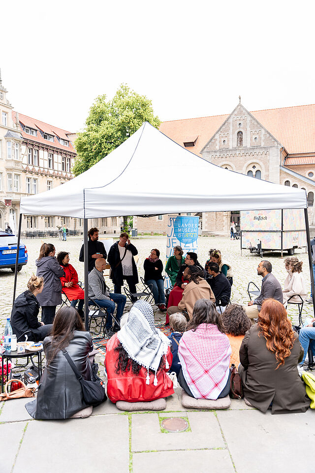 ca. 30 Personen sitzen bei einem Pavillon und hören Personen zu, die in der Mitte eine Fishbowl-Diskussion führen