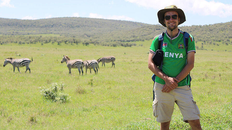 Student Nils Michalke stands in a steppe landscape. Zebras graze in the background.