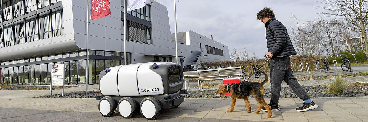 Fußgänger mit Hund und autonom fahrenden Fahrzeug vor dem NFF Gebäude. 