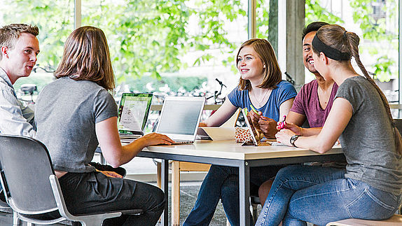 Eine Gruppe von Studierenden mit Laptops am Tisch im Gespräch